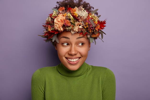 Mulher atraente e feliz tem beleza natural, não tem maquiagem, usa grinalda de planta de outono, olha alegremente de lado, sente-se satisfeita, usa casaco de malha verde, modela interior.