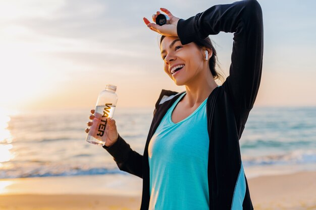 Mulher atraente e magra fazendo exercícios esportivos na praia ao nascer do sol de manhã em roupas esportivas, água potável na garrafa com sede, estilo de vida saudável, ouvindo música em fones de ouvido sem fio, dia quente de verão