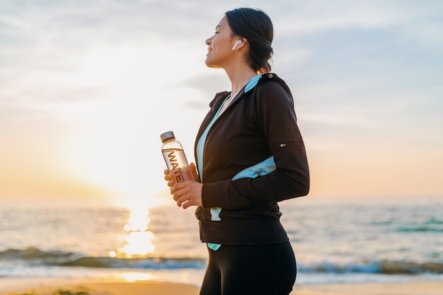 Mulher atraente e magra fazendo exercícios esportivos na praia do nascer do sol da manhã em roupas esportivas, segurando uma garrafa de água, estilo de vida saudável, ouvindo música em fones de ouvido sem fio