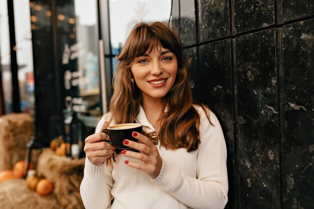 Mulher atraente e sorridente com cabelos castanhos ondulados, tomando café lá fora