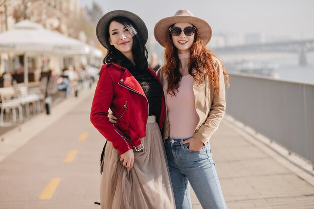 Mulher atraente e sorridente com saia bege exuberante e jaqueta vermelha, curtindo momentos de lazer com a melhor amiga