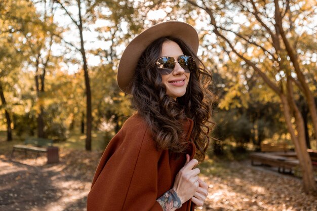 Mulher atraente, elegante e sorridente, com cabelo encaracolado andando no parque da rua, vestida com um casaco marrom quente na moda do outono, estilo de rua usando chapéu e óculos escuros