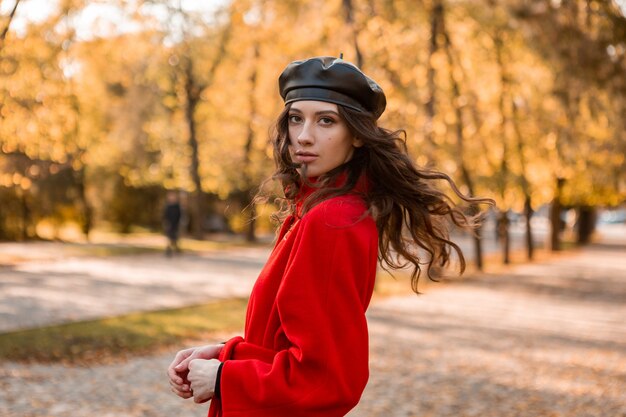 Mulher atraente, elegante e sorridente, com cabelos cacheados caminhando no parque, vestida com um casaco vermelho quente na moda do outono, estilo de rua, usando um chapéu boina