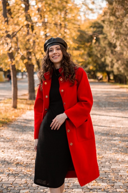 Mulher atraente, elegante e sorridente, com cabelos cacheados caminhando no parque, vestida com um casaco vermelho quente na moda do outono, estilo de rua, usando um chapéu boina