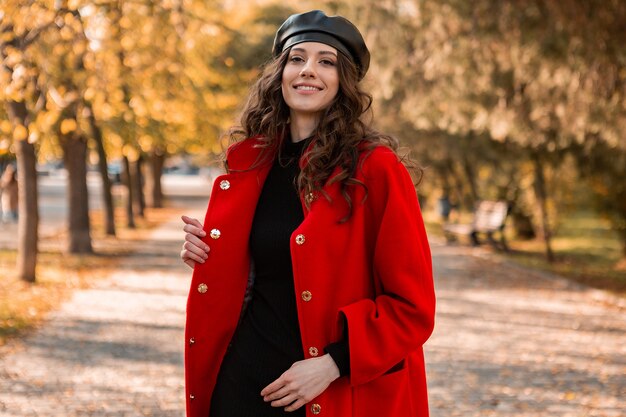 Mulher atraente, elegante e sorridente, com cabelos cacheados caminhando no parque, vestida com um casaco vermelho quente na moda do outono, estilo de rua, usando um chapéu boina