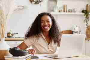 Foto grátis mulher atraente elegante jovem de pele escura em uma camisa bege, sentada à mesa da cozinha, usando o laptop, calculando o orçamento, planejando as férias, sorrindo alegremente. mulher negra autônoma trabalhando em casa