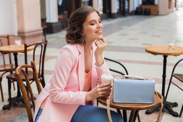 Mulher atraente em clima romântico sorrindo de felicidade sentado à mesa usando uma jaqueta rosa, roupas elegantes, esperando o namorado em um encontro no café, bebendo cappuccino, expressão facial extasiada