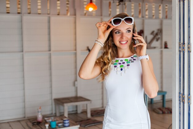 Mulher atraente em um vestido branco em um café de verão com óculos de sol falando ao telefone