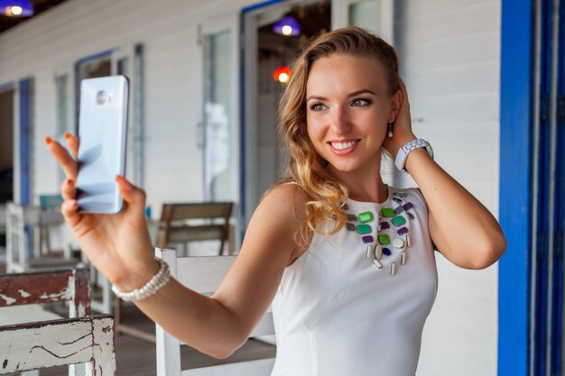 Mulher atraente em um vestido branco no verão café com óculos de sol usando telefone, tirando foto