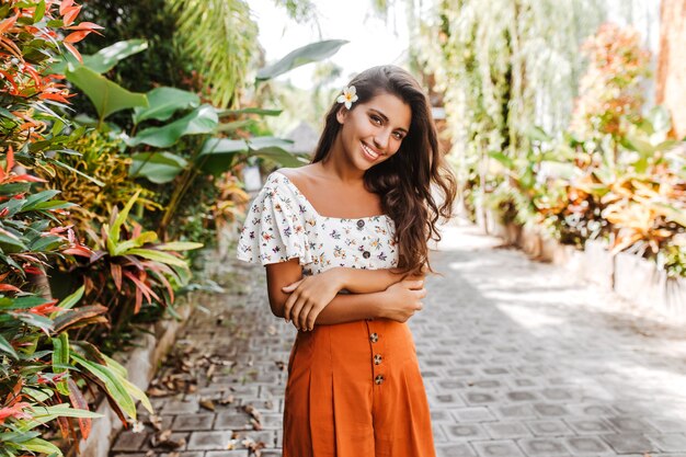 Mulher atraente encaracolada com saia laranja e blusa branca olhando para a frente com um sorriso