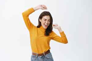 Foto grátis mulher atraente feliz dançando e se divertindo, levantando as mãos despreocupada, curtindo música, encostada na parede branca