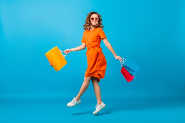 Mulher atraente feliz e elegante shopaholic em um vestido laranja superdimensionado e pulando segurando sacolas de compras no fundo azul do estúdio
