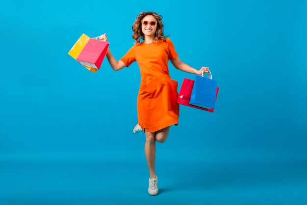 Mulher atraente feliz e elegante shopaholic em um vestido laranja superdimensionado, pulando correndo segurando sacolas de compras sobre fundo azul isoladas