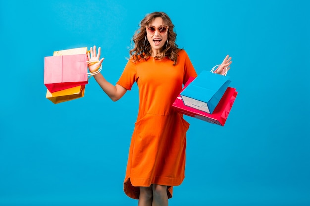 Mulher atraente feliz e elegante shopaholic em um vestido laranja superdimensionado, pulando correndo segurando sacolas de compras sobre fundo azul isoladas