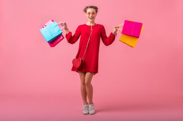 Mulher atraente, feliz e elegante, shopaholic em um vestido vermelho da moda segurando sacolas coloridas na parede rosa isolada, venda animada, tendência da moda primavera-verão