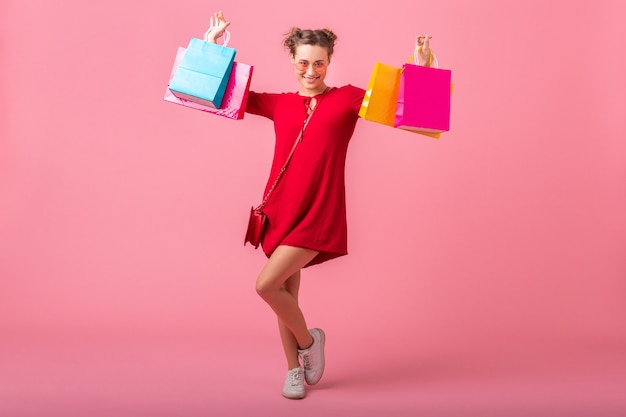 Mulher atraente, feliz e elegante, shopaholic em um vestido vermelho da moda segurando sacolas coloridas na parede rosa isolada, venda animada, tendência da moda primavera-verão