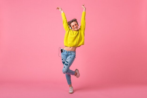 Mulher atraente feliz e engraçada em um suéter amarelo dançando ouvindo música em fones de ouvido, vestida com roupa de estilo moderno colorido isolada na parede rosa, se divertindo