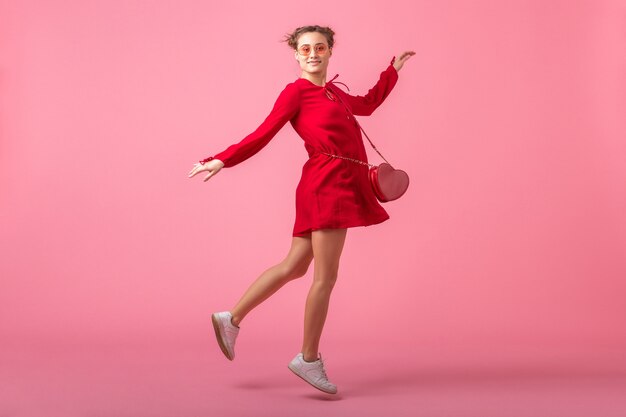 Mulher atraente, feliz e sorridente elegante em um vestido vermelho da moda, pulando correndo na parede rosa isolada, tendência da moda primavera-verão, humor romântico garota glamour