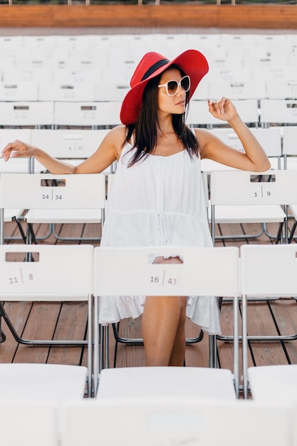 Mulher atraente feliz e sorridente vestida de vestido branco, chapéu vermelho, óculos de sol, sentada no teatro ao ar livre de verão na cadeira sozinha, tendência da moda de primavera
