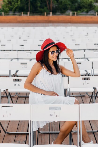 Mulher atraente feliz e sorridente vestida de vestido branco, chapéu vermelho, óculos de sol, sentada no teatro ao ar livre de verão na cadeira sozinha, tendência da moda de primavera