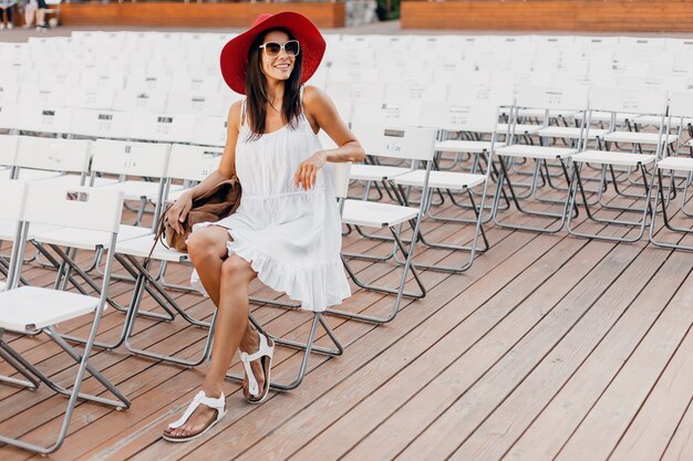 Mulher atraente feliz e sorridente vestida de vestido branco, chapéu vermelho, óculos escuros, sentada no teatro ao ar livre de verão sozinha, muitas cadeiras, tendência da moda de estilo de rua de primavera, acenando com a mão.