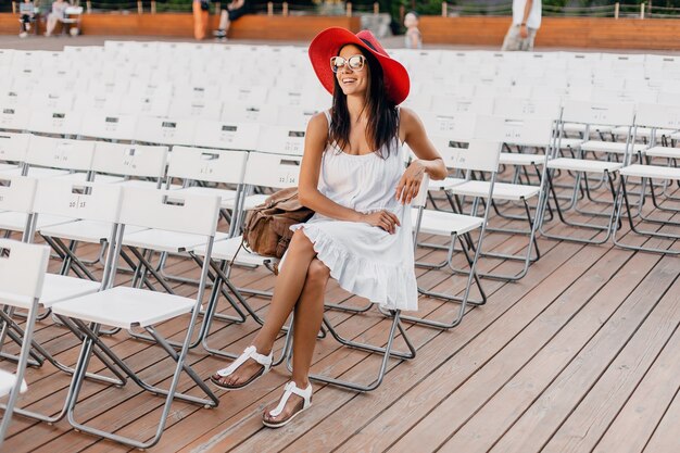 Mulher atraente feliz e sorridente vestida de vestido branco, chapéu vermelho, óculos escuros, sentada no teatro ao ar livre de verão sozinha, muitas cadeiras, tendência da moda de estilo de rua de primavera, acenando com a mão.