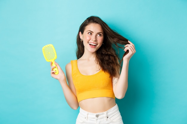 Mulher atraente feliz mostrando a escova sem fio de cabelo e tocando o penteado longo saudável com rosto satisfeito, sorrindo encantado com o resultado de shampoo, fundo azul.