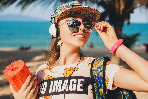 Mulher atraente hippie caminhando na praia ouvindo música em fones de ouvido em uma roupa elegante e descolada em férias de verão tropical ensolarado usando acessórios boné óculos, sorrindo feliz segurando tapete de ioga