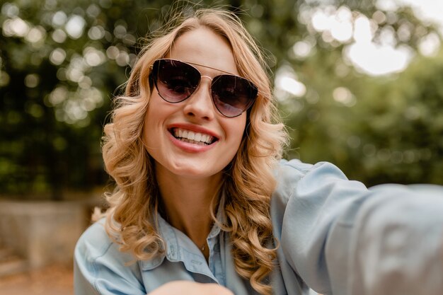 Mulher atraente loira sorridente de dentes brancos andando no parque com roupa de verão tirando foto de selfie no telefone