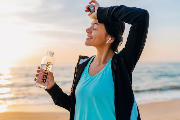 Foto grátis mulher atraente magro fazendo exercícios de esporte na praia ao nascer do sol de manhã em roupas esportivas, água potável na garrafa com sede, estilo de vida saudável, ouvindo música em fones de ouvido sem fio, sorrindo feliz