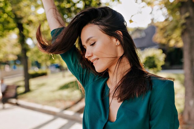 Mulher atraente morena alegre de vestido verde com sorriso maravilhoso passa o dente ao ar livre olha para baixo e posa do lado de fora