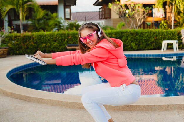 Mulher atraente praticando esportes na piscina com capuz rosa colorido com óculos escuros, ouvindo música em fones de ouvido nas férias de verão, jogar tênis, estilo esportivo
