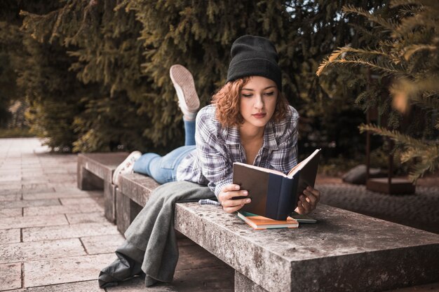 Mulher atraente ruiva no chapéu deitado no banco com livro