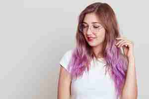 Foto grátis mulher atraente sonhadora com cabelo lilás olhando para longe e tocando seu cabelo, usando óculos e camiseta branca, copie o espaço para propaganda, posando isolado sobre uma parede cinza.