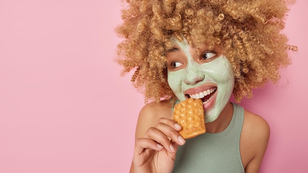 Foto grátis mulher bastante positiva com cabelo espesso encaracolado morde delicioso waffle aplica máscara de beleza no rosto para tratamento de pele passa por procedimentos antienvelhecimento usa camiseta casual isolada sobre parede rosa