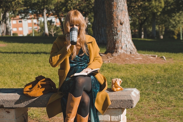 Mulher bebendo e lendo livro no parque