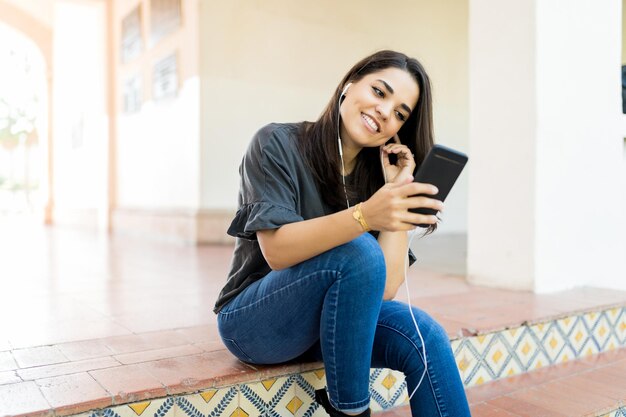 Mulher bonita adulta média ouvindo transmissão de rádio via aplicativo móvel enquanto está sentado na entrada