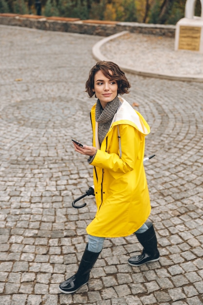 Mulher bonita andando no sett pelo parque da cidade com telefone celular e guarda-chuva nas mãos, aproveitando o dia de folga