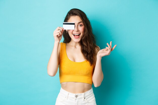 Mulher bonita animada em um top cortado de verão, olha maravilhada com a câmera, fazendo compras com cartão de crédito, em pé sobre um fundo azul