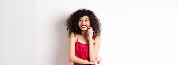 Foto grátis mulher bonita animada em vestido vermelho mordendo o dedo e sorrindo fofa para a câmera quer algo em pé