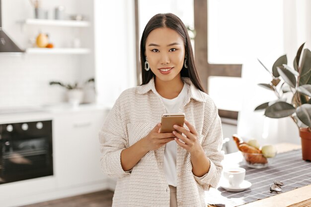 Mulher bonita asiática em um cardigã bege e camiseta branca posa com o telefone na cozinha