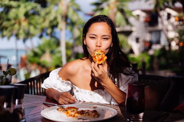Mulher bonita asiática feliz com fome comendo pizza no dia de sol luz do sol no restaurante ao ar livre Feminino desfrutando de comida se divertindo na hora do almoço