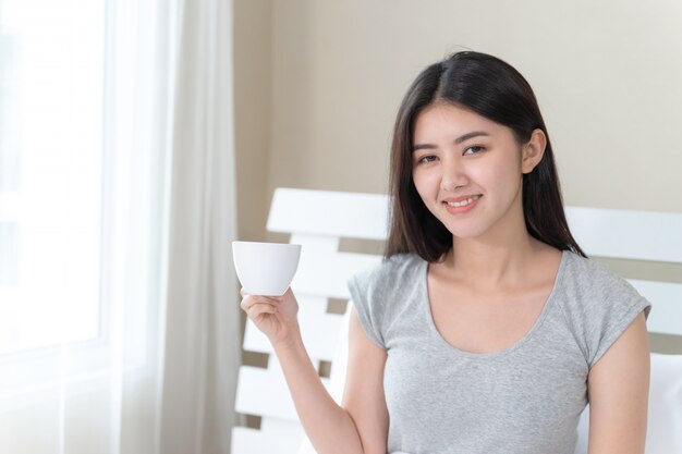 Mulher bonita asiática, sentada na cama no quarto e segurando a xícara de café na mão com feliz