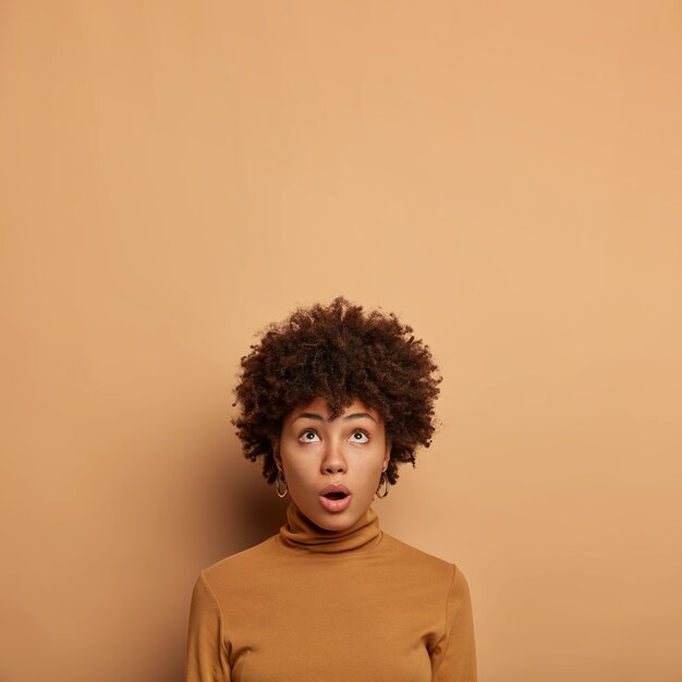 Mulher bonita atônita encara com expressão omg acima, deixa cair o queixo de espanto, usa decote de polonês marrom, concentrada no teto em grande descrença