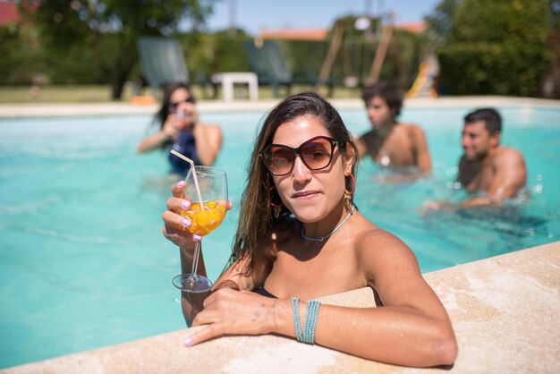 Mulher bonita bebendo um cocktail brilhante na piscina. Mulher com cabelo escuro, segurando um copo com uma bebida brilhante, olhando para a câmera. Lazer, amizade, conceito de festa