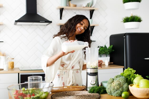 Mulher bonita biracial sorrindo está derramando leite no liquidificador perto da mesa com legumes frescos na cozinha branca moderna vestida com pijamas e cabelos soltos