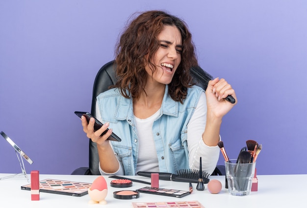 Mulher bonita caucasiana alegre sentada à mesa com ferramentas de maquiagem segurando o telefone e o pente fingindo cantar isolado na parede roxa com espaço de cópia
