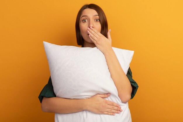 Mulher bonita caucasiana ansiosa segurando um travesseiro e colocando a mão na boca em uma laranja