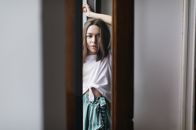 Mulher bonita caucasiana com expressão de rosto calmo, relaxando em seu apartamento. Magnífica menina morena com roupas casuais, posando em casa.