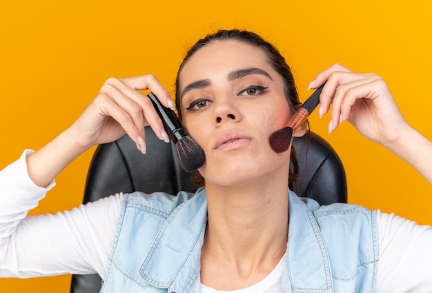 Mulher bonita caucasiana confiante sentada à mesa com ferramentas de maquiagem, aplicando blush com pincéis de maquiagem isolados na parede laranja com espaço de cópia